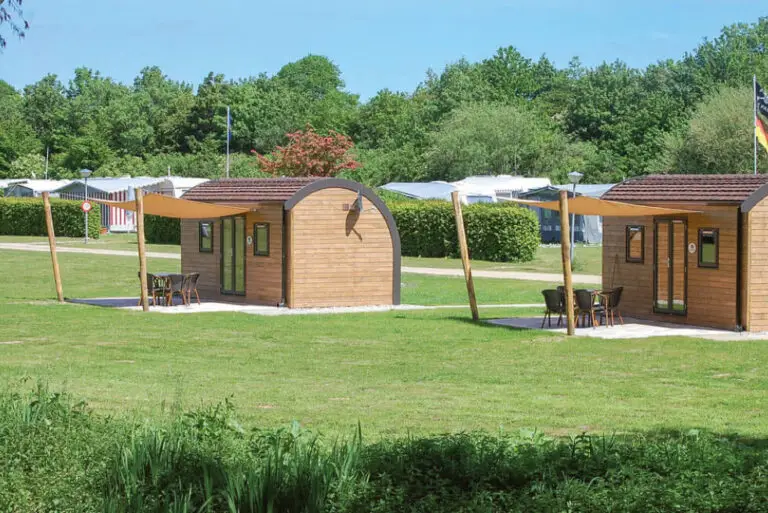 Zwei SeeLodges im Sonnenlicht auf der grünen Rasenfläche des Campingplatzes mit Holzwänden, einem runden Dach, 2 kleinen Fenstern und eine doppelflügelige Glastür, davor jeweills eine gepflasterte Terrasse mit Tisch, vier Stühlen und Sonnensegel