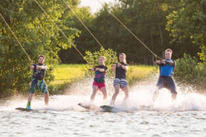 Süsel Seeparx - 4 vier Wakeboarder gleichzeitig nebeneinander auf dem Wasser mit grüner Wiese und baumbewachsenen Ufer im Hintergrund