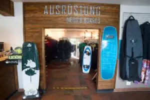 Süsel Seeparx Wakeboard Store Eingang mit Wakeboards und zugehörigen Taschen, Blick durch den Eingang auf Kleiderstangen mit Neoprenanzügen