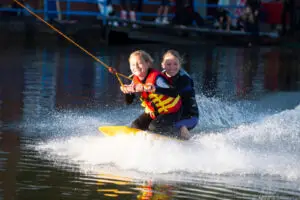 Zwei strahlende Mädchen knien auf dem Kneeboard bei voller Fahrt und spritzendem Wasser