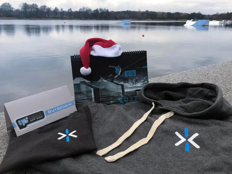 graues T-Shirt, grauer Hoodie, ein Gutschein und der Kalender 2022 von Süsel Seeparx liegen vor der Kulisse des Sees in Süsel